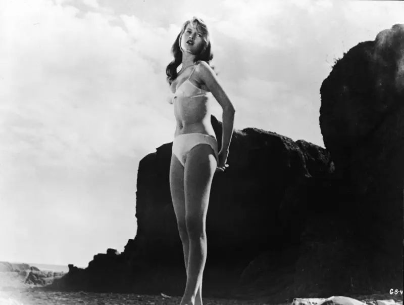 Bardot wears a white bikini and stands on a rocky beach in a still from The Girl in the Bikini (1958), directed by Willy Rozier — Hulton Archive @ Getty Image